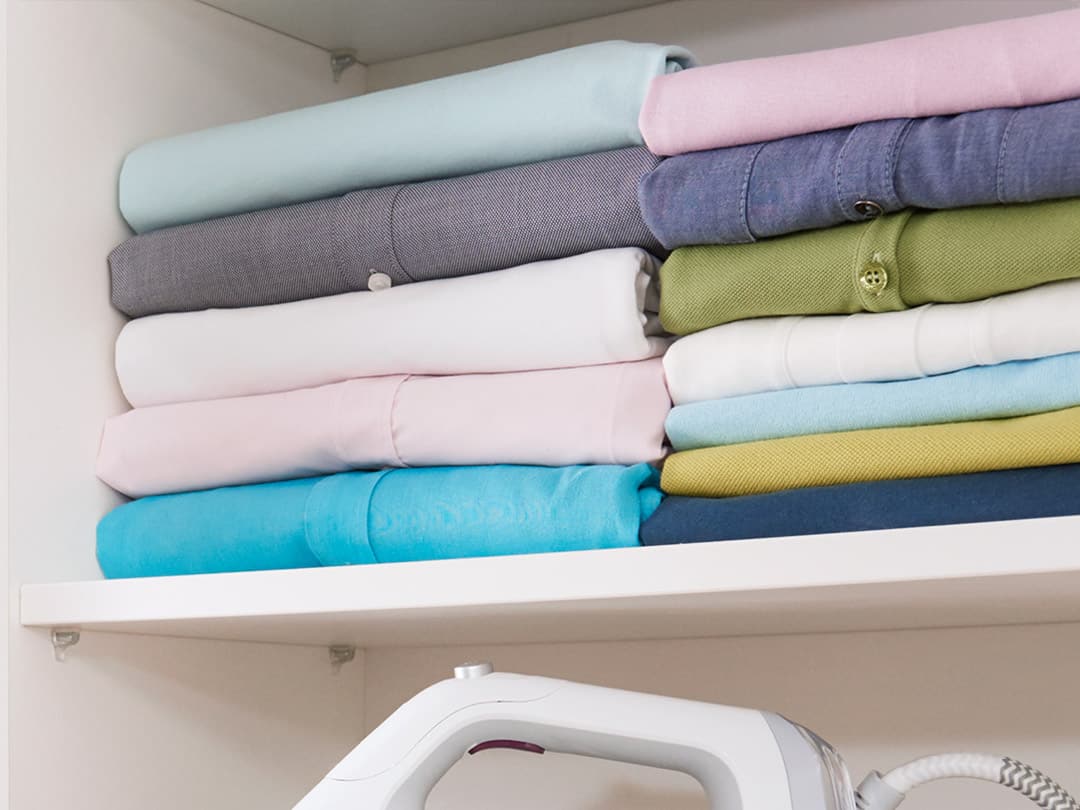 A white shelf with an ironing board and folded clothes.