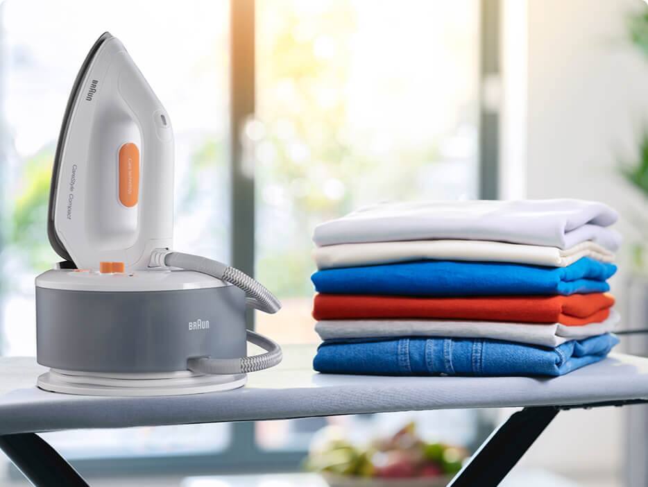 A Braun CareStyle Compact steam generator iron in its station on an ironing board with a pile of freshly folded laundry.
